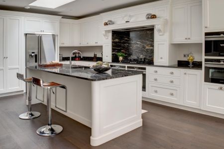 A traditional bespoke kitchen with white panelled cabinets, black marble countertops, a central island with seating, dark wood flooring, and a striking stone backsplash.