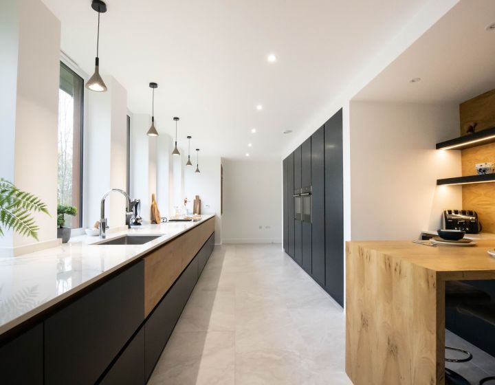 A modern bespoke kitchen with long, dark grey cabinets, white countertops, natural wood open shelving, and a wood-topped breakfast bar, illuminated by unique pendant lights and complemented by pale tiled flooring.