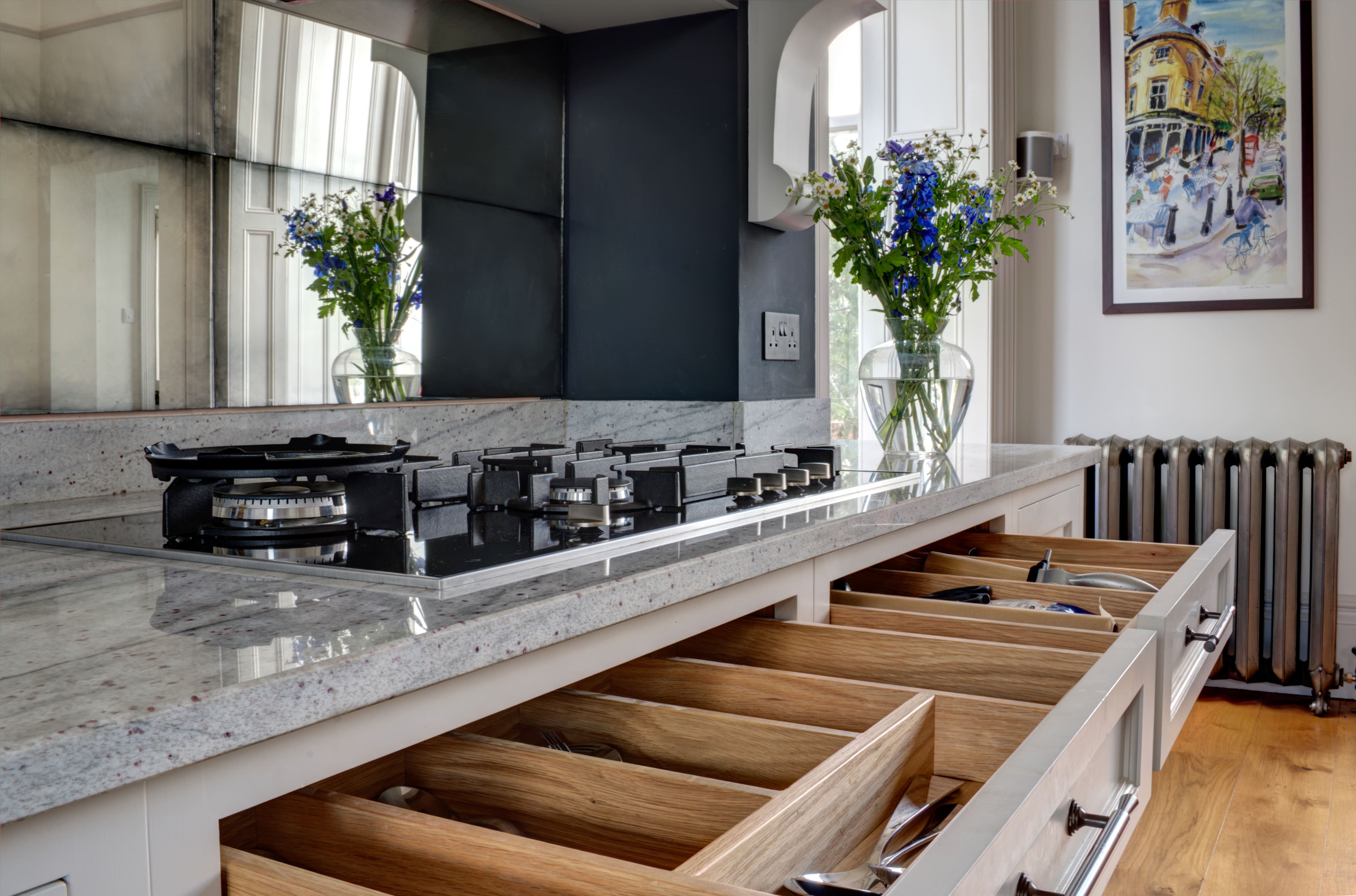 A modern kitchen detail showing a gas stove top integrated into a granite countertop with a reflective black splashback. In the foreground, several wooden drawers are pulled open, revealing neatly organised kitchen utensils. 