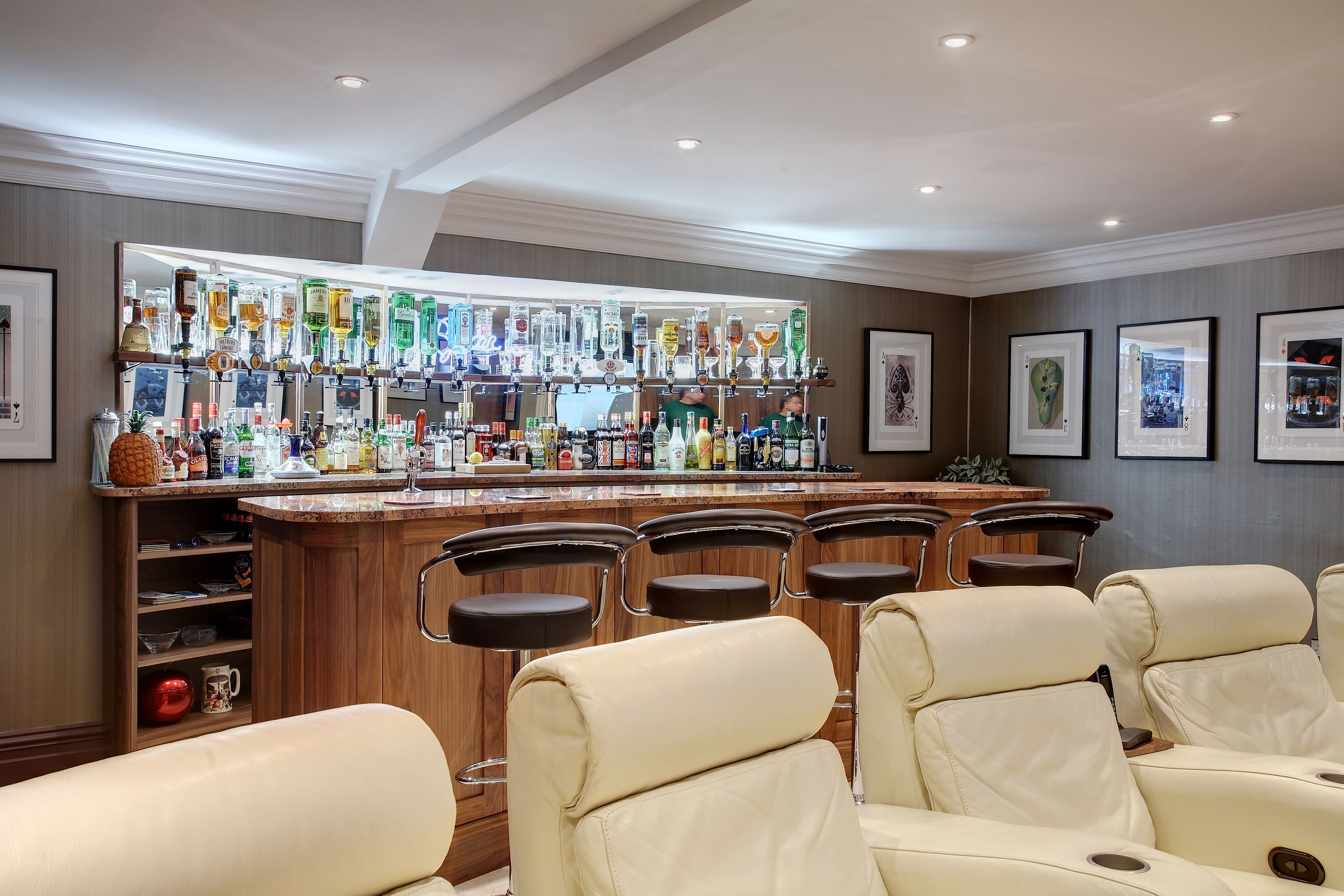 A large, bespoke personal bar. A number of bottles are backlit, leading down to a varnished wood bar. Some large cream-coloured leather chairs sit in the foreground.