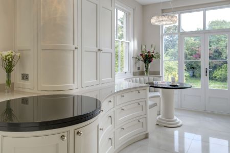 A cream-coloured bespoke Shaker kitchen, with a circular seating area with a round black table looks out some external doors to a colourful garden.