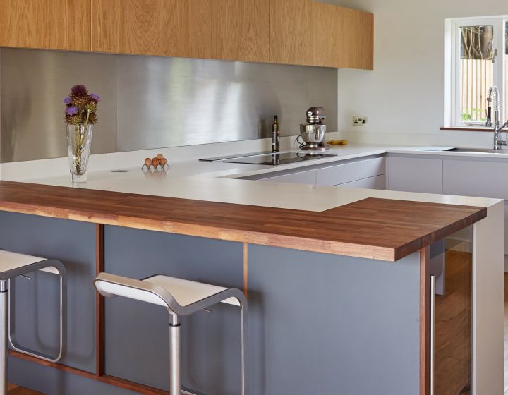 A sleek bespoke kitchen with charcoal grey cabinets, walnut worktops, a modern induction hob, stainless steel backsplash, and light oak flooring, accented by contemporary bar stools and pendant lighting.