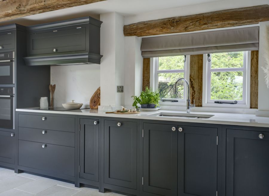 A series of dark blue, bespoke kitchen cabinets with white worktops strongly contrast some overhead, rustic oak beams. A small window over integrated-sink looks to some greenery.