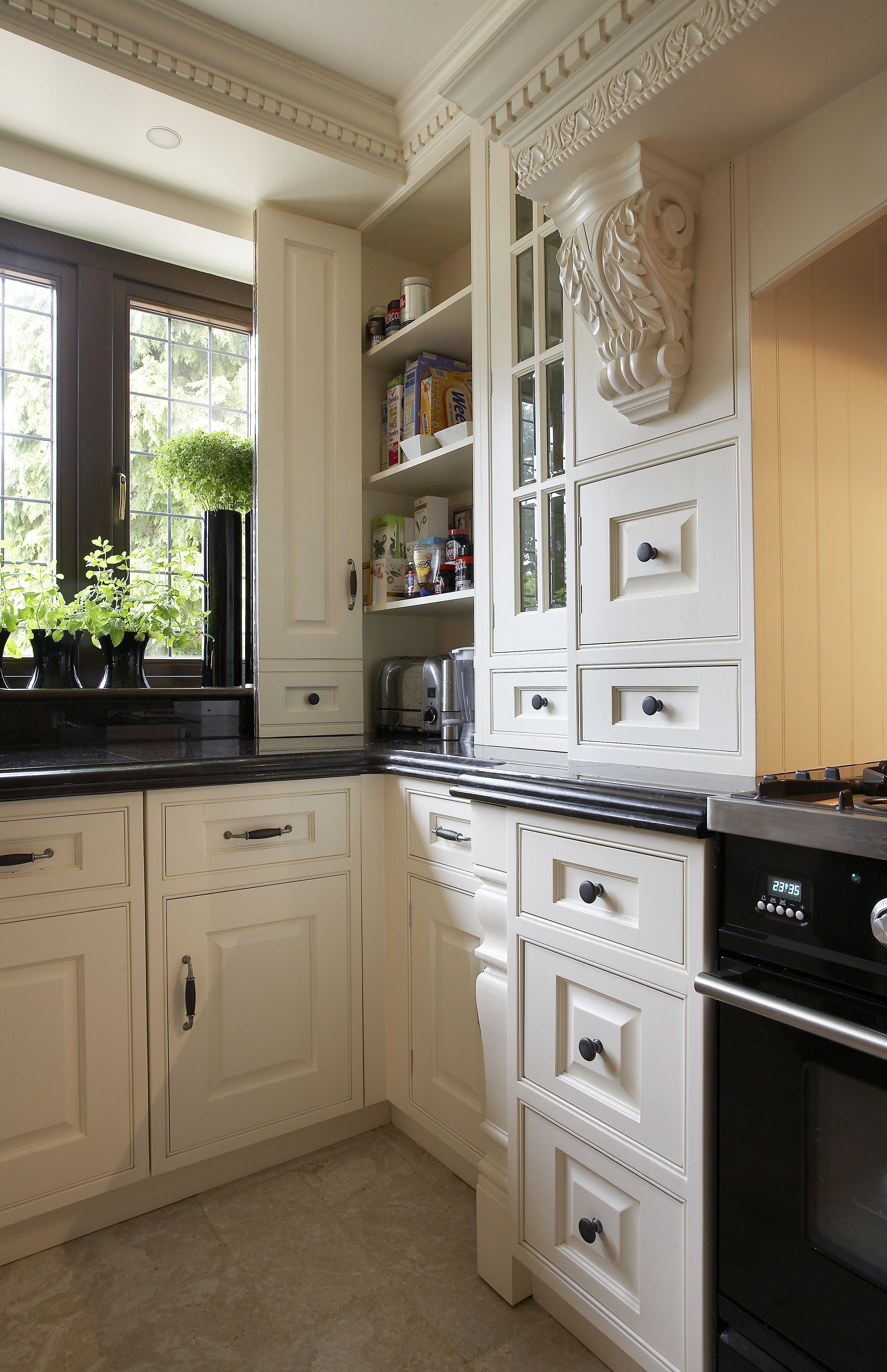  A traditional kitchen corner with classic cream-colored cabinetry featuring intricate cornice and corbel details. The cabinets are topped with a dark granite countertop. 