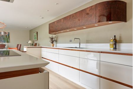 A modern bespoke kitchen with white high-gloss cabinetry, walnut accents, white countertops, and a unique burl wood extractor hood, all complemented by warm oak flooring and subtle recessed lighting.