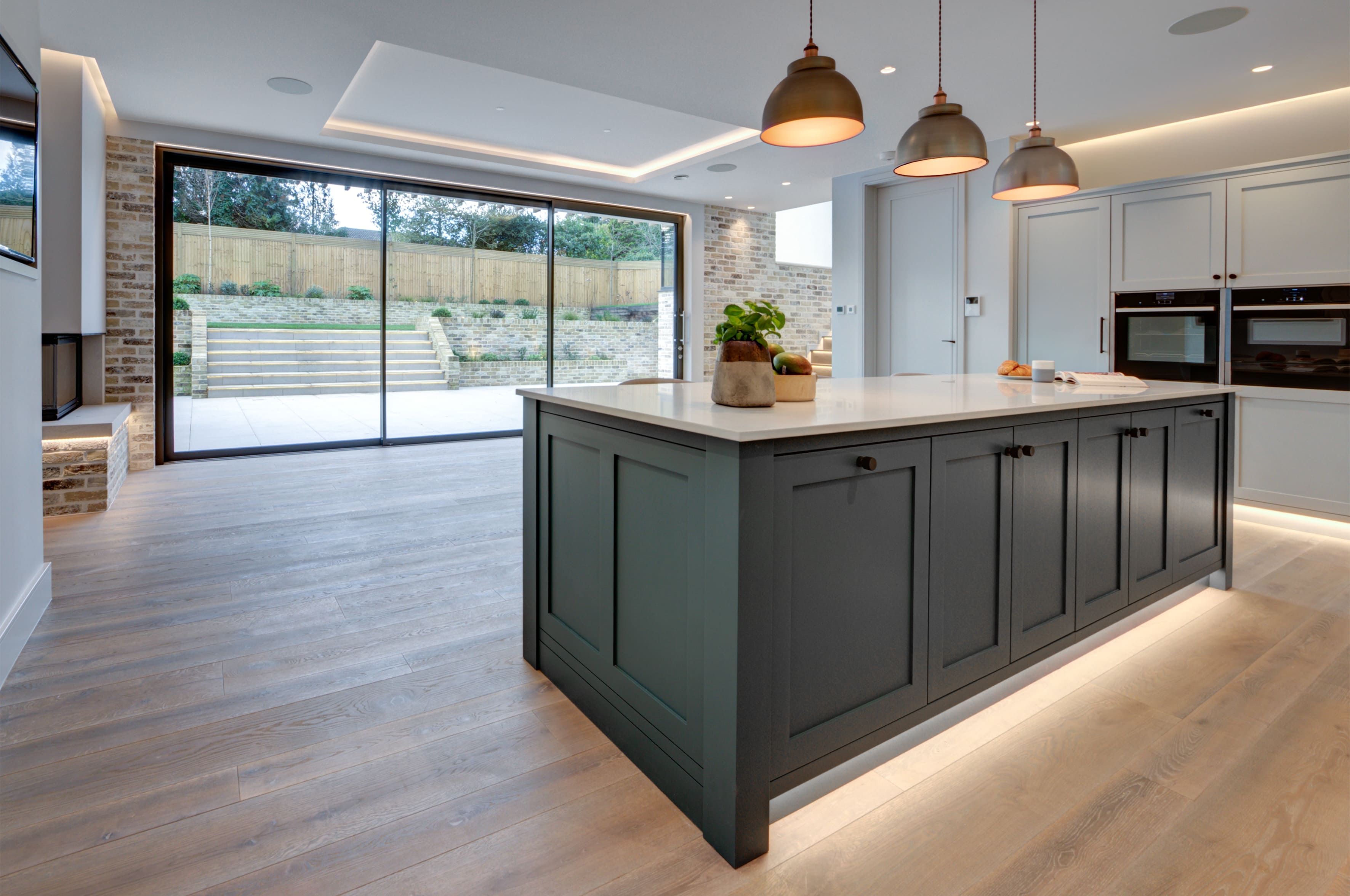 A large room with wooden tiled flooring features a large, bespoke kitchen island with under-counter lighting. Large floor-to-ceiling patio doors flood the room with natural light.