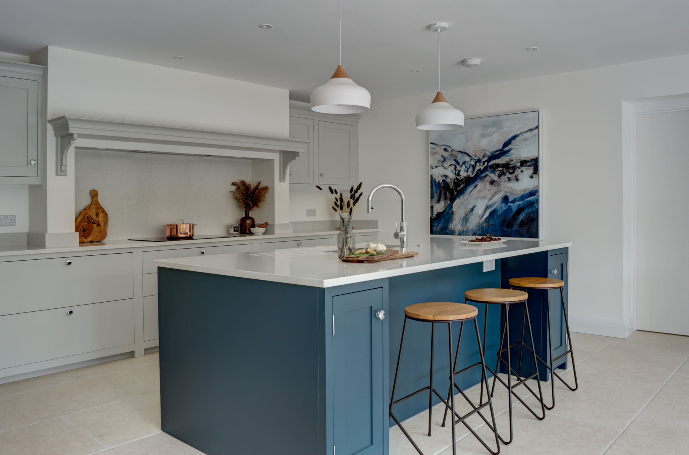 A contemporary bespoke kitchen with a two-tone design, featuring white cabinets, a teal island with a white countertop, wooden bar stools, pendant lighting, and decorative art on the wall, set on a light tiled floor.