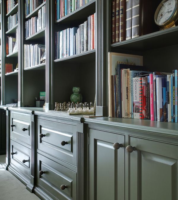 A sophisticated home study with dark green built-in bookshelves filled with a colorful array of books. In front of the bookshelves is a classic chest of drawers with a traditional design and dark metal handles.