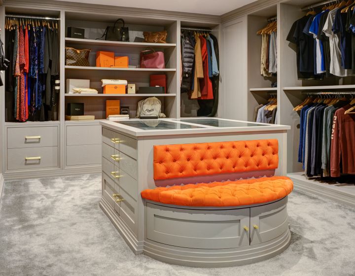 A bespoke dressing room with integrated cabinets surrounding the room. In the centre is a counter, with a striking orange-cushioned ottoman.