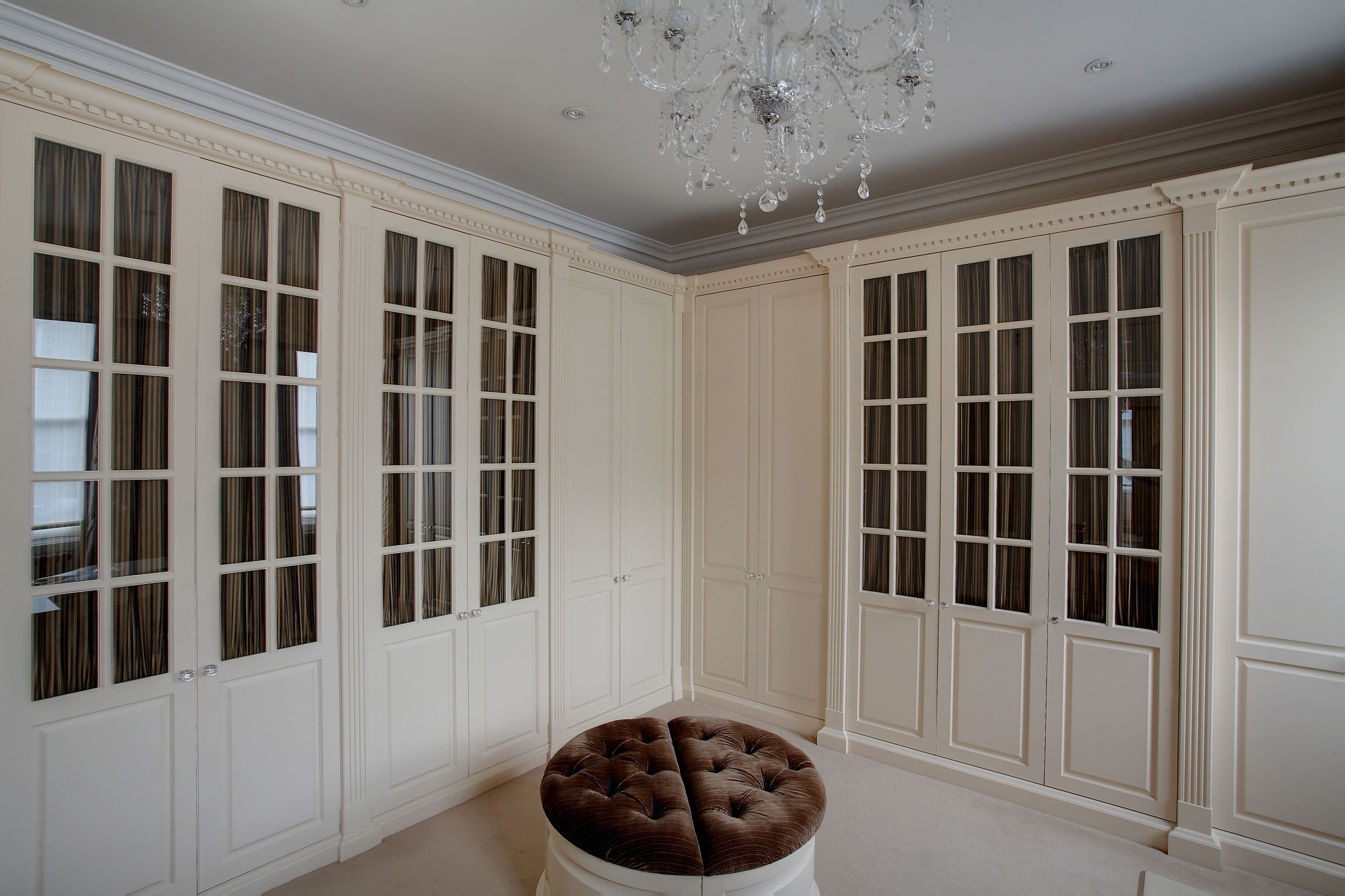 A luxurious bedroom with a neutral colour palette, featuring a large, circular tufted ottoman in the centre. The room has cream fitted wardrobes with paneled doors and glass inserts, offering plenty of storage space. An elaborate crystal chandelier hangs from the ceiling, adding a touch of elegance.