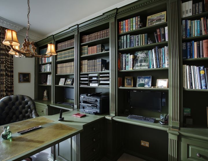 A sophisticated home study with dark green built-in bookshelves filled with a colourful array of books. In front of the bookshelves is a classic chest of drawers with a traditional design and dark metal handles.