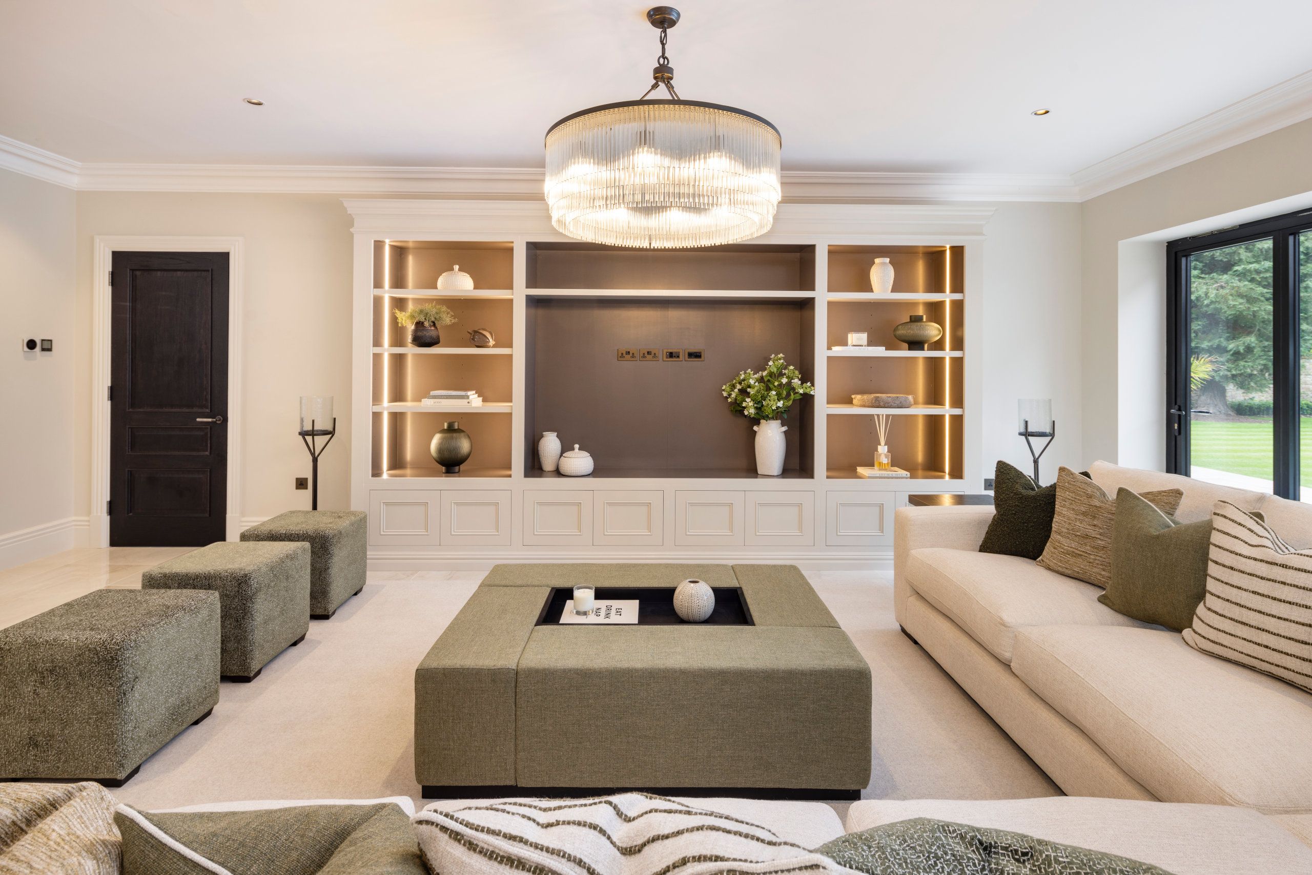 A luxurious cinema room with a contemporary design. The room features a large, cream-colored L-shaped sofa adorned with various textured throw pillows. In the centre is a large square ottoman used as a coffee table.