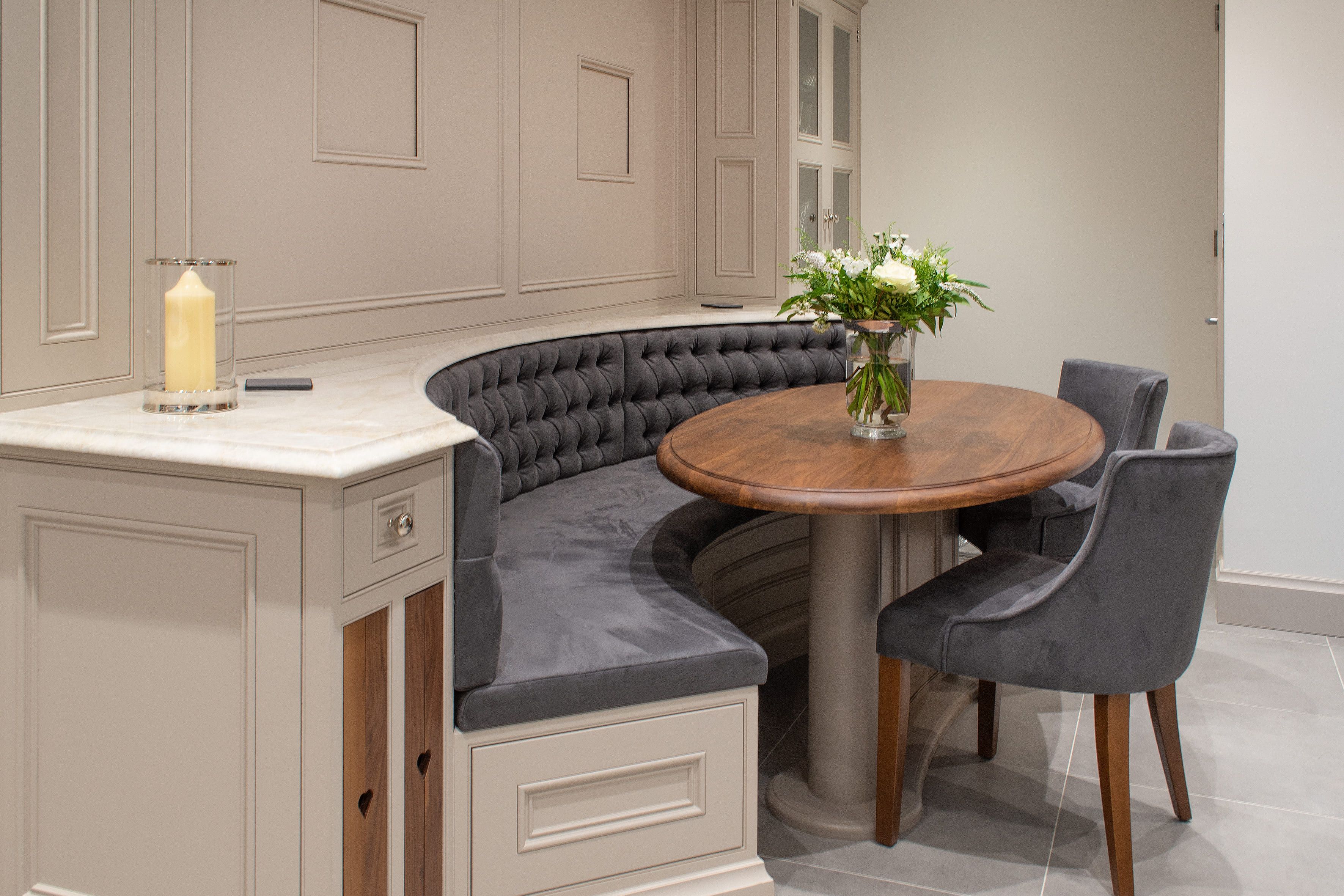 A cozy bespoke kitchen nook with a built-in bench upholstered in dark grey tufted fabric and two matching chairs. The round wooden table complements the bench and chairs. A vase with fresh flowers is placed at the centre of the table. 