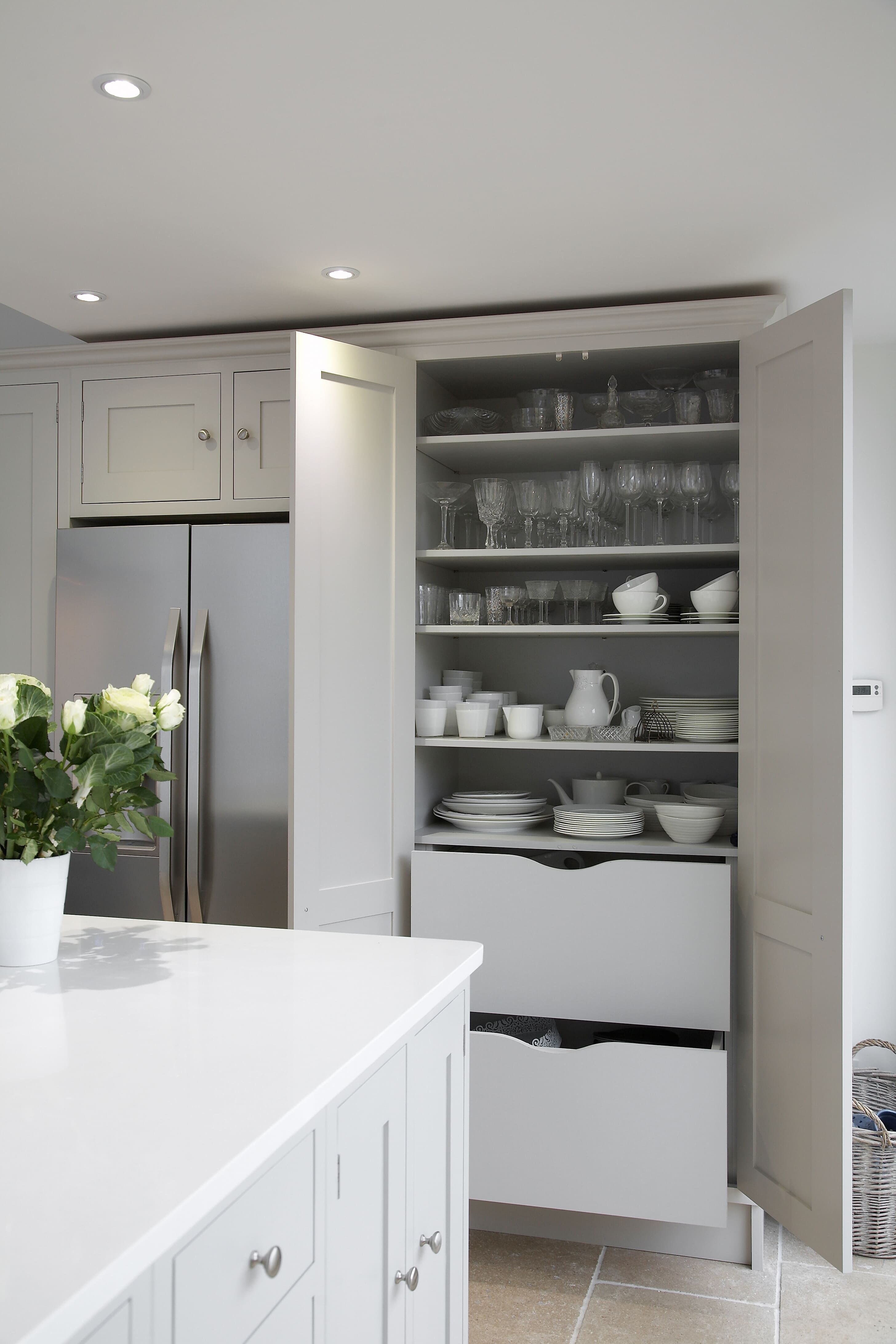 A modern kitchen pantry open to reveal neatly organised shelves with glassware, dishes, and bowls. The cabinetry is painted in a soft grey, and the handles are a classic style in brushed metal. 